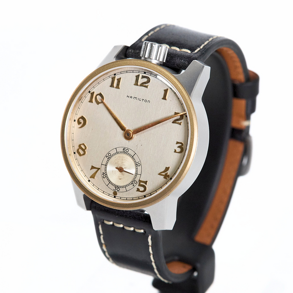 A vintage wristwatch with a round silver-toned case, gold-tone bezel and hands, a cream-colored dial with black Arabic numerals, and a black leather strap with white stitching and a brown interior lining, shown on a plain white background.