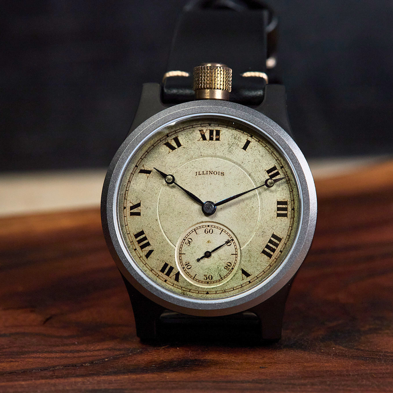 A vintage-style wristwatch with a round silver-toned case, cream dial, black strap, and gold-tone crown, photographed standing on a wooden surface against a dark blurred background.