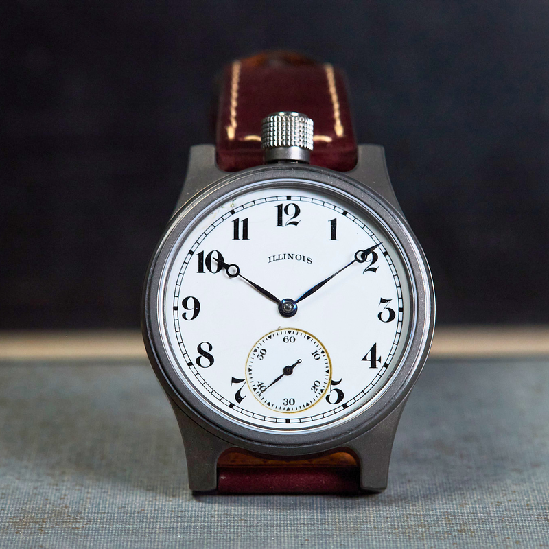 An Illinois wristwatch with a round silver-tone case, white dial, black Arabic numerals, and a brown leather strap, photographed standing upright against a dark background.