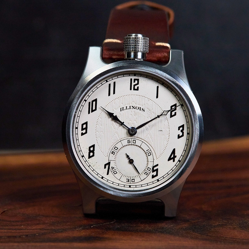 An Illinois brand analog watch with a silver-toned metal case, white dial with black Arabic numerals, and a brown leather strap, resting on a dark wooden surface against a dark background.