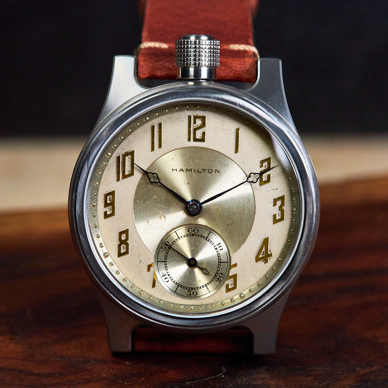 A vintage-style Hamilton wristwatch with a round silver-toned case, cream dial, black Arabic numerals, and a brown leather strap, photographed close up on a wooden surface.