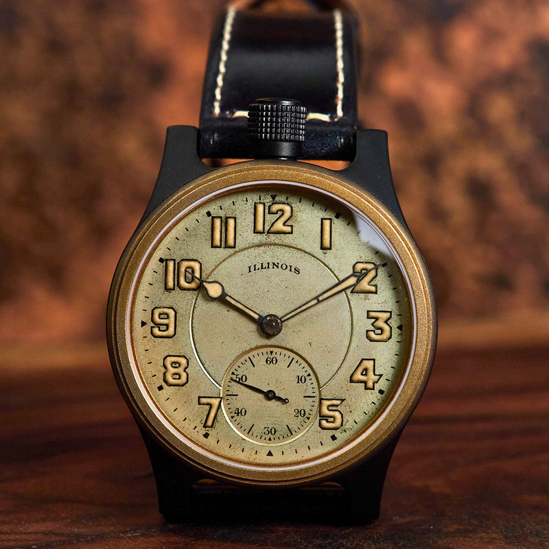 A vintage-style wristwatch with a round black case, gold-tone bezel, cream dial with black Arabic numerals, and a black leather strap with white stitching, photographed on a wooden surface against a warm brown background.
