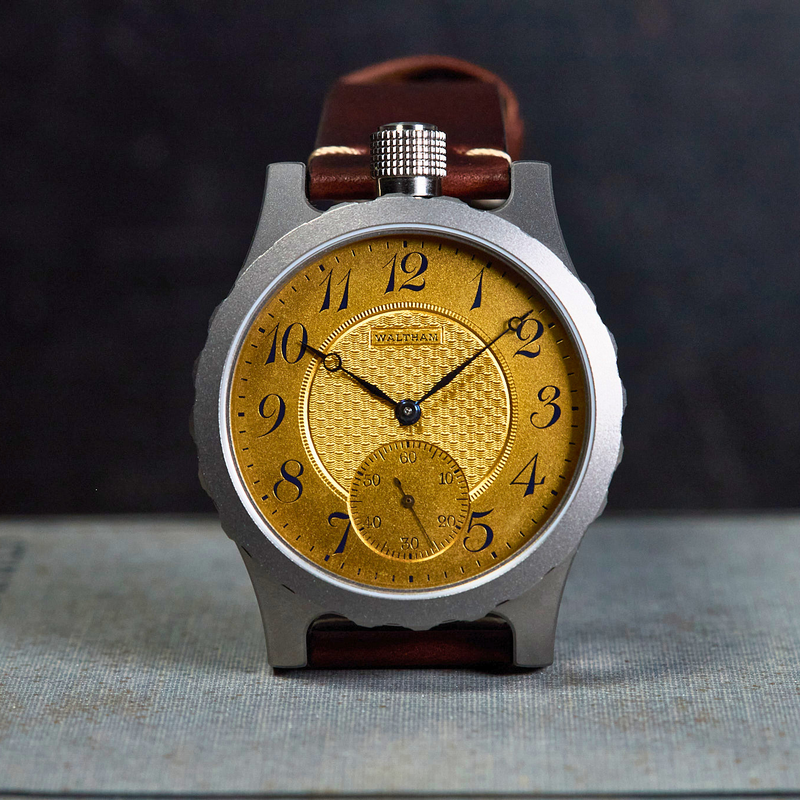 A wristwatch with a textured gold dial, black Arabic numerals, and a silver-toned case, shown standing upright on a textured surface against a dark background.