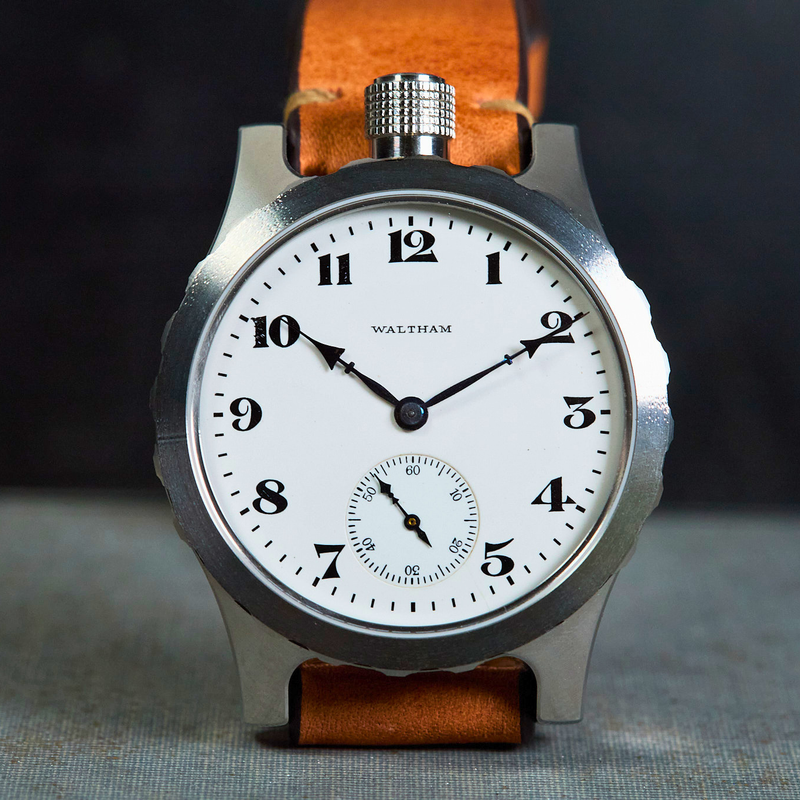 A Waltham watch with a silver-toned metal case, white dial, and brown leather strap, photographed upright against a dark background with a textured surface below.