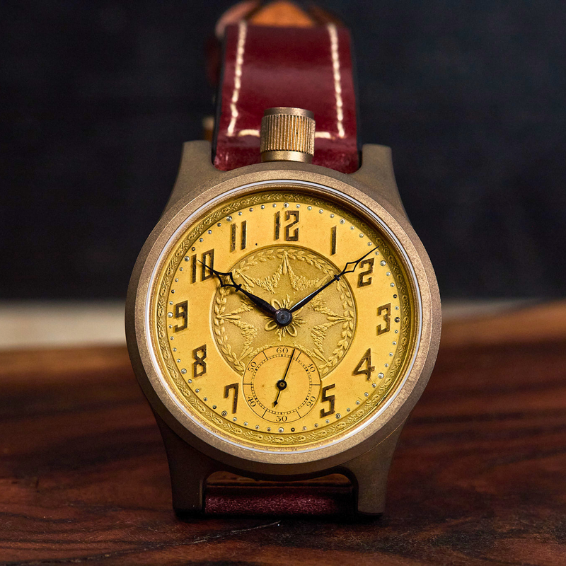 A vintage-style wristwatch with a round bronze-toned case, a gold dial featuring black Arabic numerals and a small subdial, and a dark reddish-brown leather strap, shown standing on a wooden surface against a dark background.