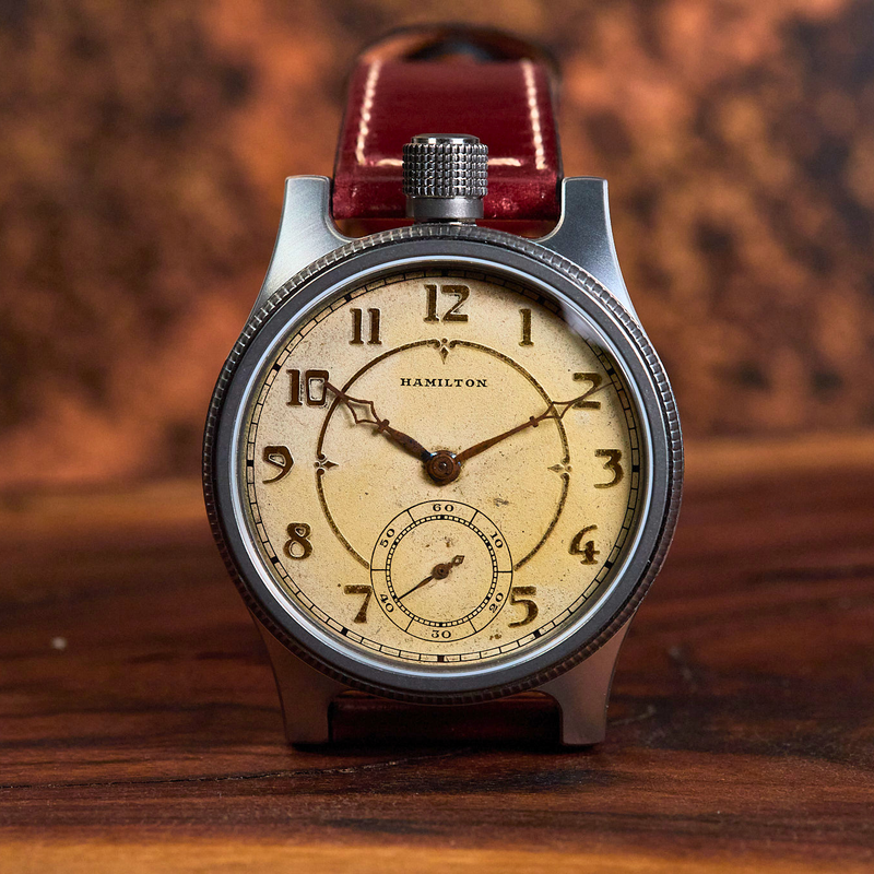 A Hamilton wristwatch with a silver-toned case, cream dial, and red leather strap, displayed on a wooden surface with a blurred warm brown background.