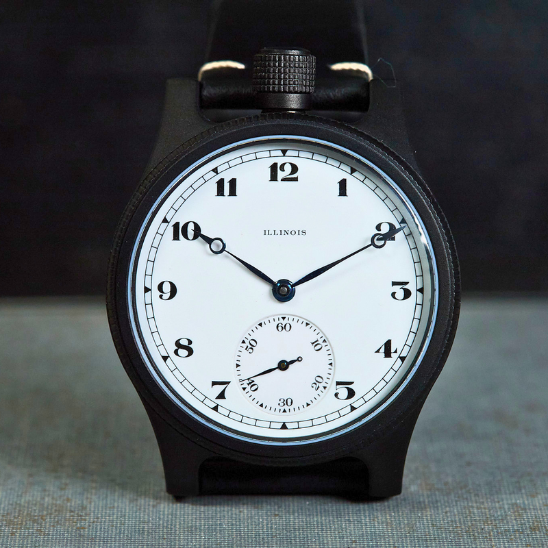 A black dial watch with a white face, black Arabic numerals, and a black strap, photographed upright against a dark background with a textured surface below.