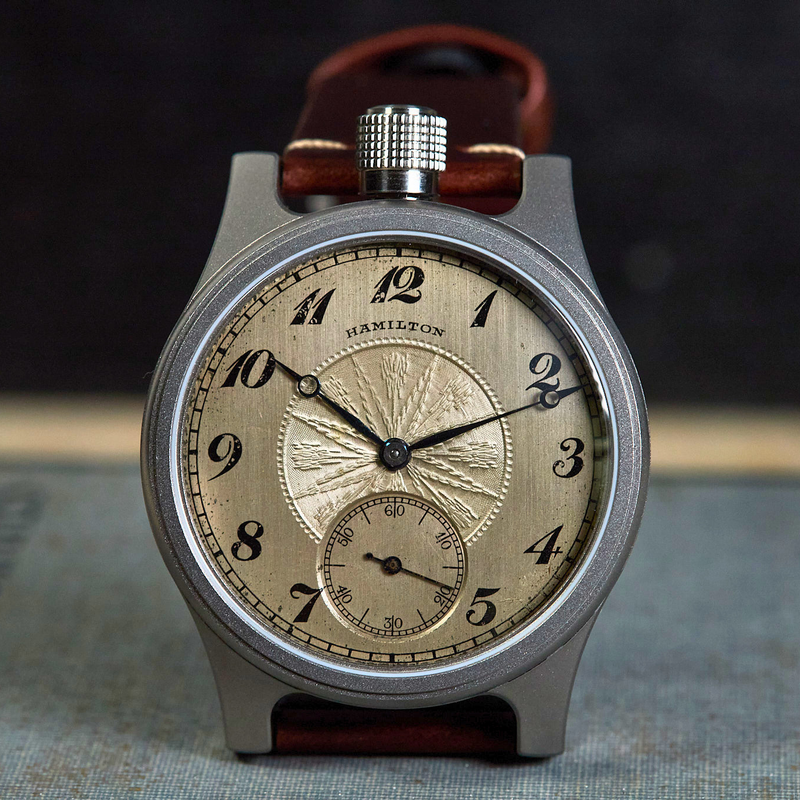 A vintage-style wristwatch with a round silver-toned case, a textured beige dial featuring black Arabic numerals and a small subdial, and a brown leather strap, photographed upright against a dark blurred background.