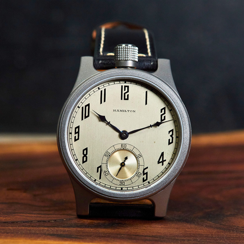 A Hamilton watch with a round silver-toned case, cream-colored dial, and black leather strap, displayed on a wooden surface against a dark background.