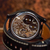 Close-up of a black smartwatch with a transparent back showing a detailed mechanical movement, attached to a brown leather strap on a wooden surface.