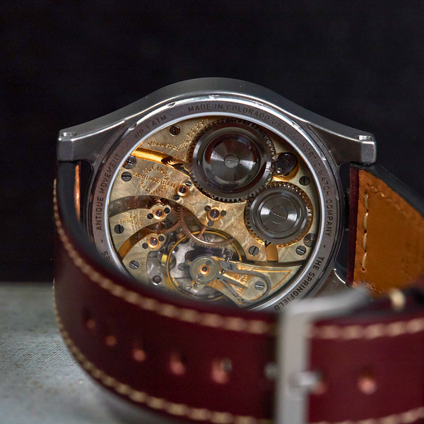A close-up shot of a skeleton watch showing its intricate gold-toned mechanical movement through a dark brown leather strap with white stitching.