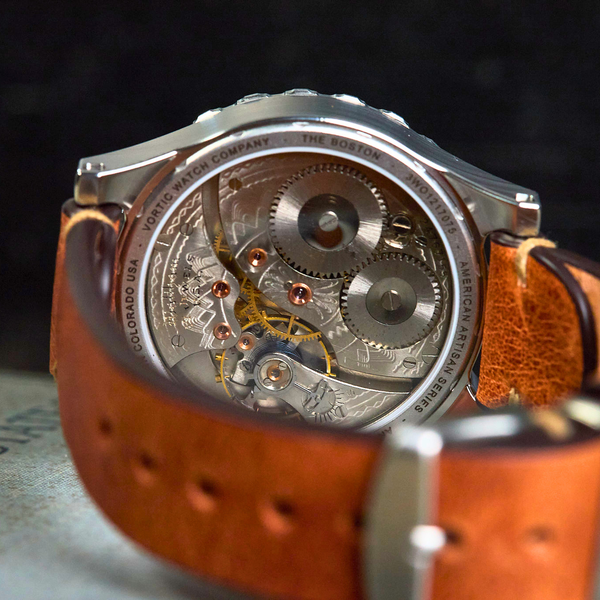 A close-up shot of a silver-toned automatic watch with a transparent case showing the intricate mechanical movement inside, paired with a brown leather strap.