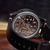A close-up of a black wristwatch with a transparent dial showing a detailed mechanical movement, resting on a wooden surface against a dark background.