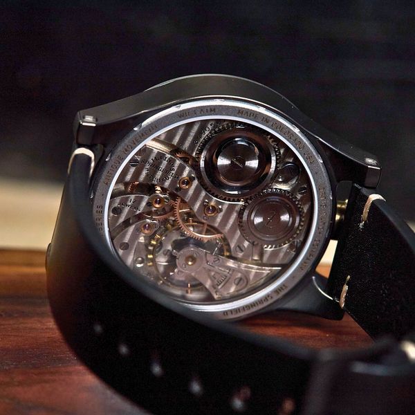 A close-up of a black wristwatch with a transparent dial showing a detailed mechanical movement, resting on a wooden surface against a dark background.