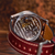 A close-up shot of a luxury watch with a silver-toned case, showing the intricate mechanical movement through a transparent backplate, attached to a dark red leather strap on a wooden surface.