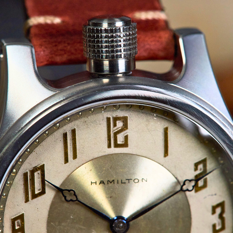 Close-up of a vintage-style wristwatch with a silver-toned metal case, cream-colored dial, black numerals, and a reddish-brown leather strap. The brand name HAMILTON is visible on the dial.