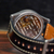Close-up of a wristwatch with a transparent back showing a detailed mechanical movement, paired with a dark brown leather strap and a tan leather section, resting on a wooden surface.