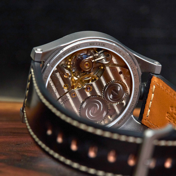 Close-up of a wristwatch with a transparent back showing a detailed mechanical movement, paired with a dark brown leather strap and a tan leather section, resting on a wooden surface.