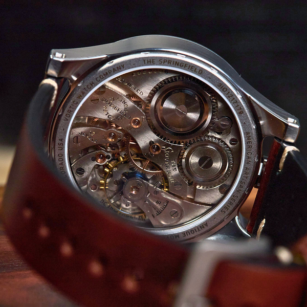 Close-up of a wristwatch with a transparent back showing a detailed mechanical movement, paired with a brown leather strap on a wooden surface.