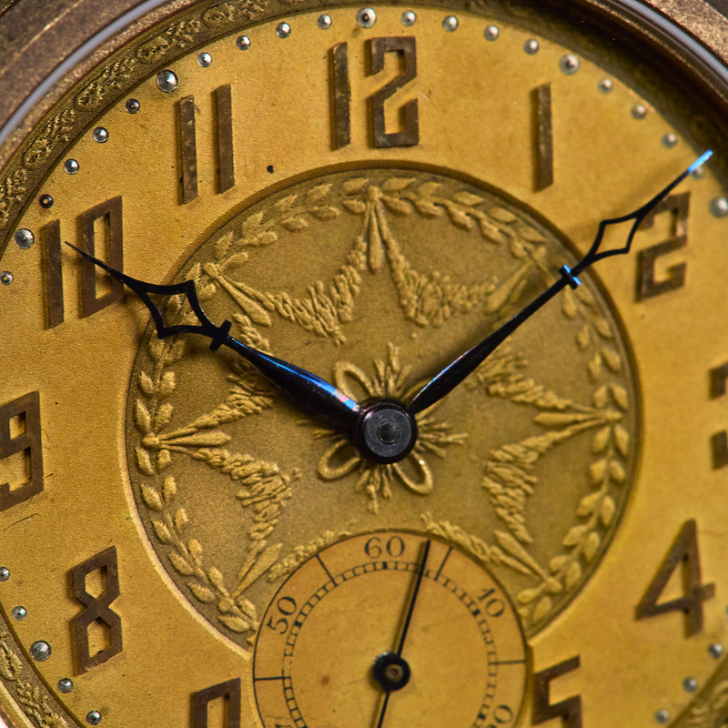 Close-up of a vintage-style watch dial with a gold-toned face, black Arabic numerals, ornate decorative detailing, and black hands.