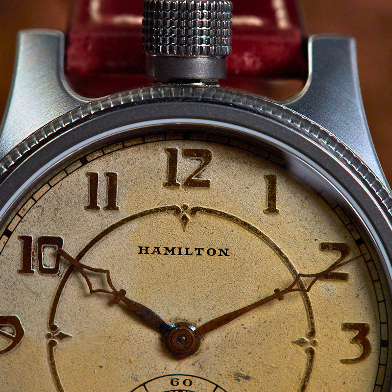 Close-up of a vintage Hamilton watch with a textured silver-tone case, a cream-colored dial with dark numerals, and a red leather strap visible at the top.