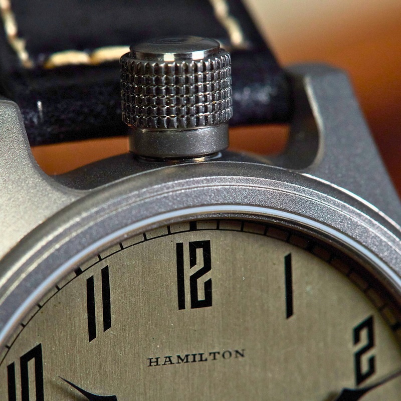 Close-up of a Hamilton watch with a textured metal case, black dial numerals, and a dark leather strap against a warm blurred background.