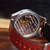 A close-up shot of a mechanical watch from the back, showcasing the detailed silver-toned movement through a transparent case, attached to a brown leather strap resting on a wooden surface.