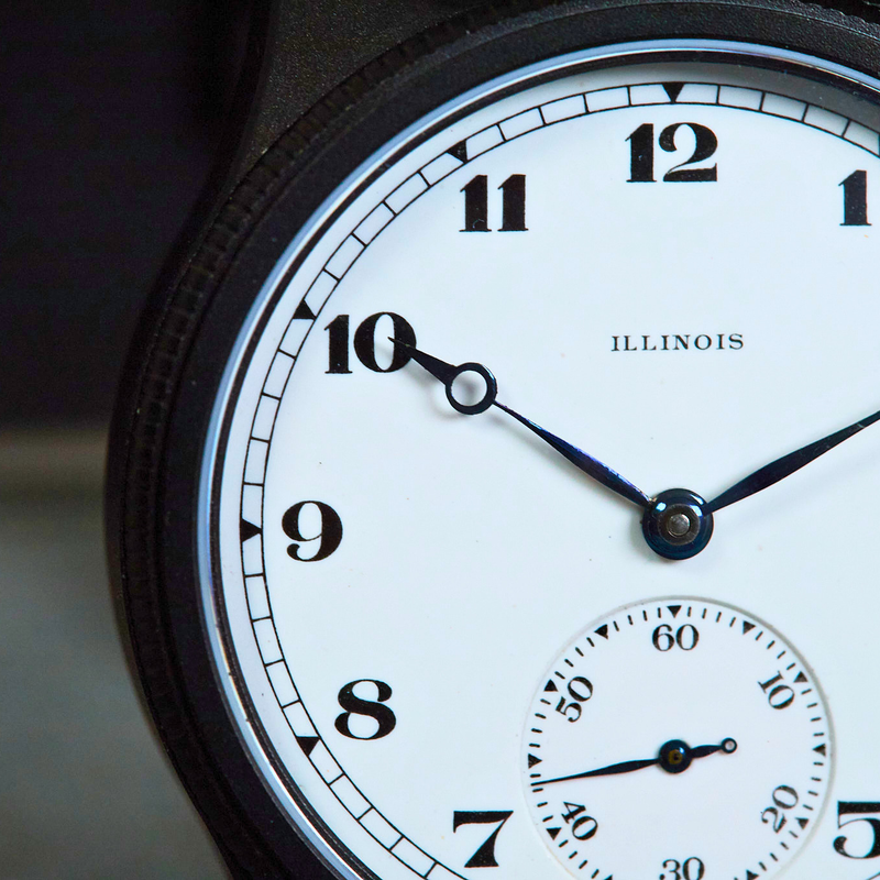 Close-up of a black watch face with a white dial, black Arabic numerals, and a small subdial at the bottom. The word 'ILLINOIS' is printed on the dial.