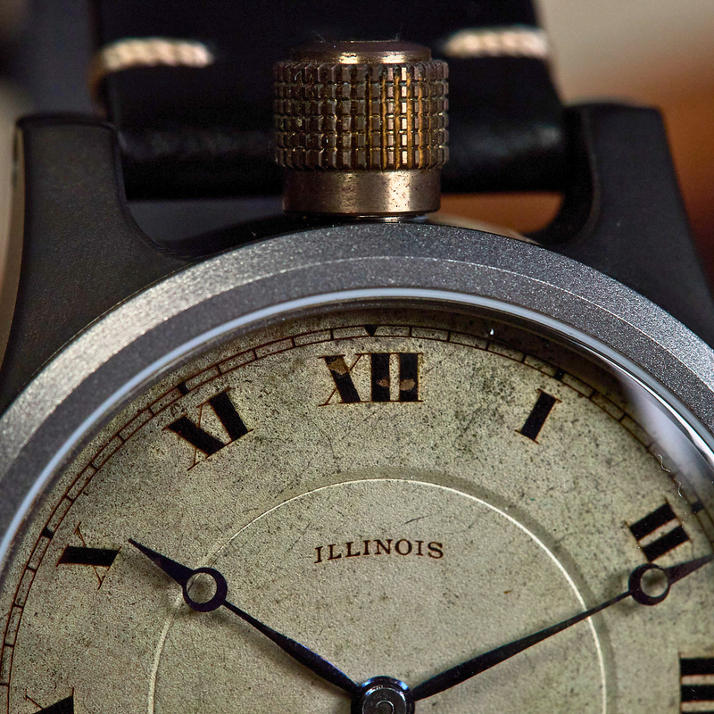 Close-up of a vintage-style wristwatch with a round silver-toned case, distressed cream dial, black Roman numerals, and a black strap with white stitching. The word 'ILLINOIS' is printed on the dial.