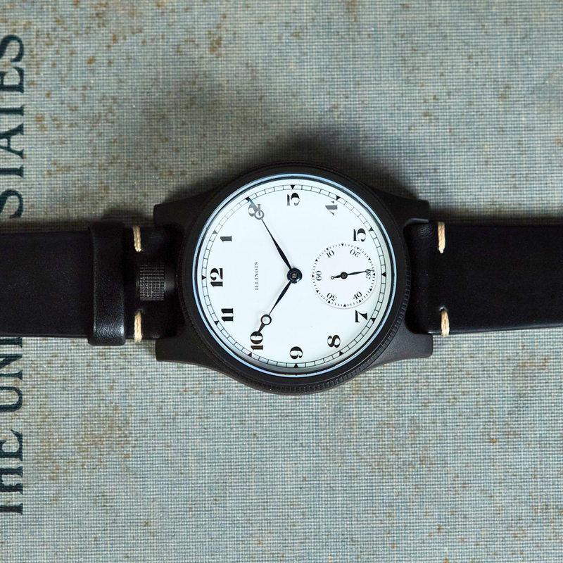 A vintage-style wristwatch with a black leather strap and a black case, featuring a white dial with black Arabic numerals and hands, laid flat on a textured gray surface with printed text.