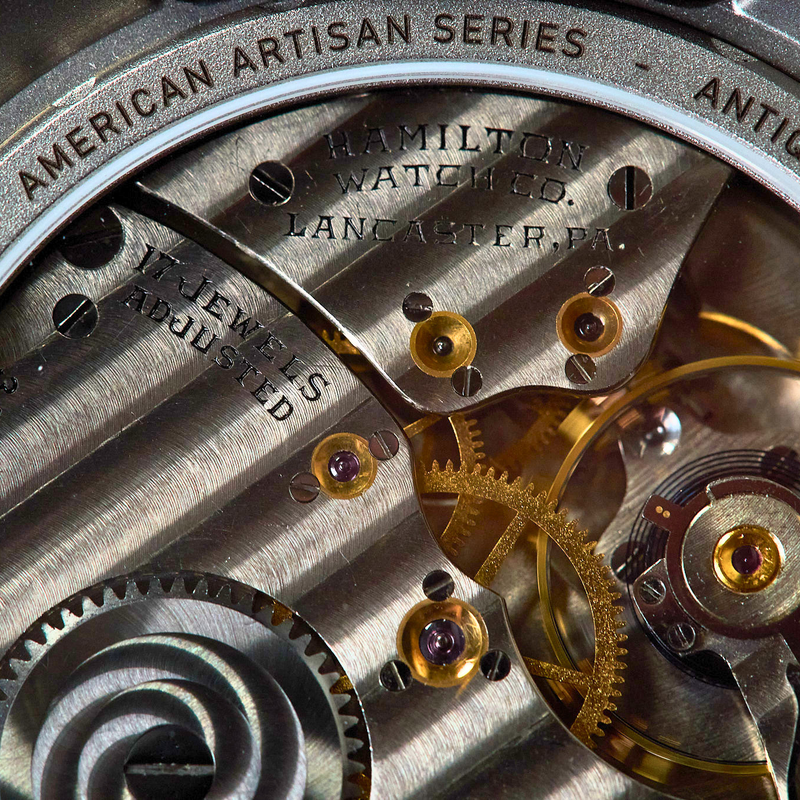 Close-up of a Hamilton Watch Co. mechanical watch movement, showing gears, screws, and engraved text including 'American Artisan Series' and 'Antique Movement'.