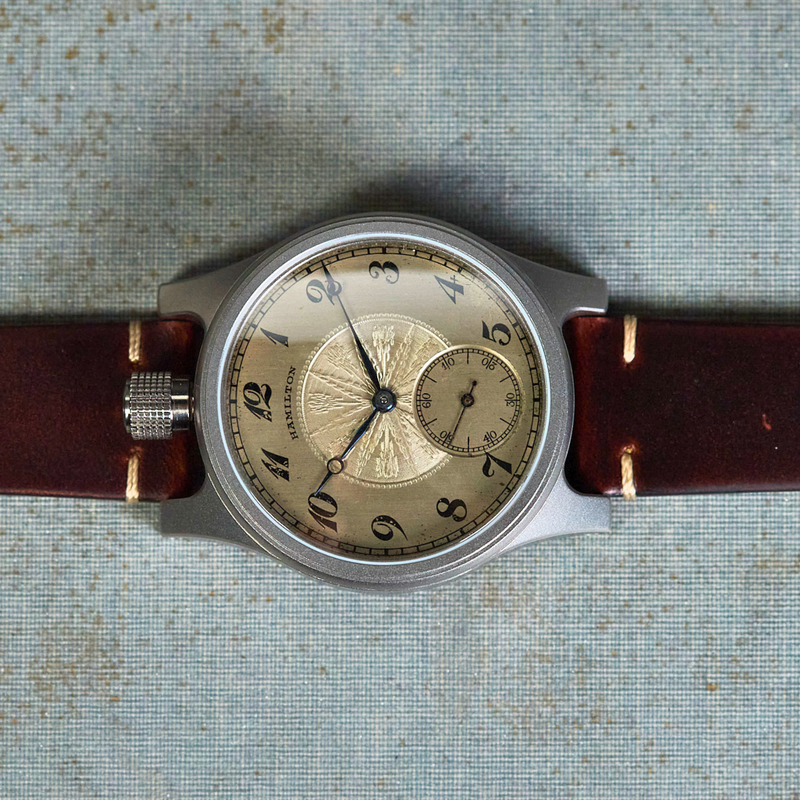 A vintage-style wristwatch with a round silver-toned case, beige sunburst dial, and dark brown leather strap, laid flat on a textured light blue surface with printed text.