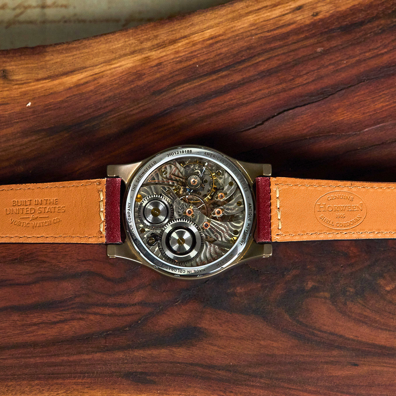 A skeleton watch with a round silver-toned case, revealing intricate internal gears, paired with a tan leather strap and burgundy stitching, laid flat on a dark wood background.