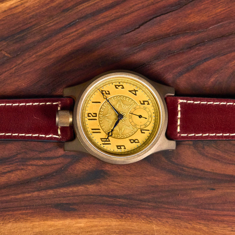 A wristwatch with a round gold dial, black hands, and a dark red leather strap with white stitching, laid flat on a dark wood background.