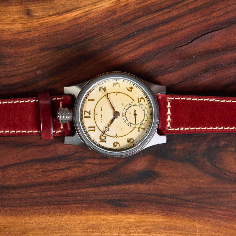 A vintage-style wristwatch with a cream dial, silver-toned case, and a deep red leather strap, laid flat on a dark wood background.