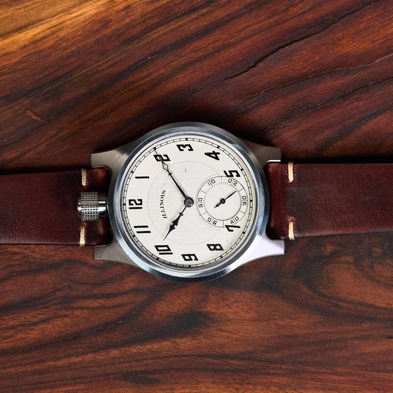 A vintage-style wristwatch with a brown leather strap and a silver-toned case, laid flat on a dark wood surface. The watch has a white dial with black Arabic numerals and a small seconds subdial.