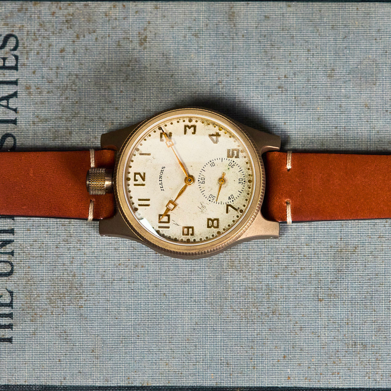 A vintage-style wristwatch with a round cream dial, gold-tone case, and brown leather strap, laid flat on a textured blue-gray book cover with printed text.