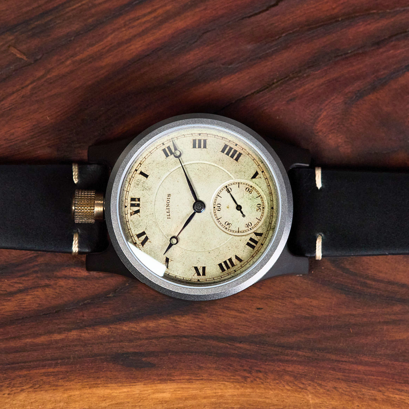 A vintage-style wristwatch with a black leather strap, round silver-toned case, and cream dial with black Roman numerals, photographed on a dark wood surface.