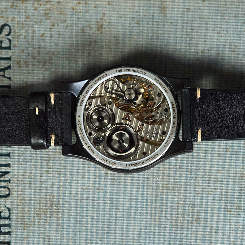 A close-up of a skeleton watch showing the intricate mechanical movement through a black leather strap, laid flat on a textured light blue-gray surface with vertical text.
