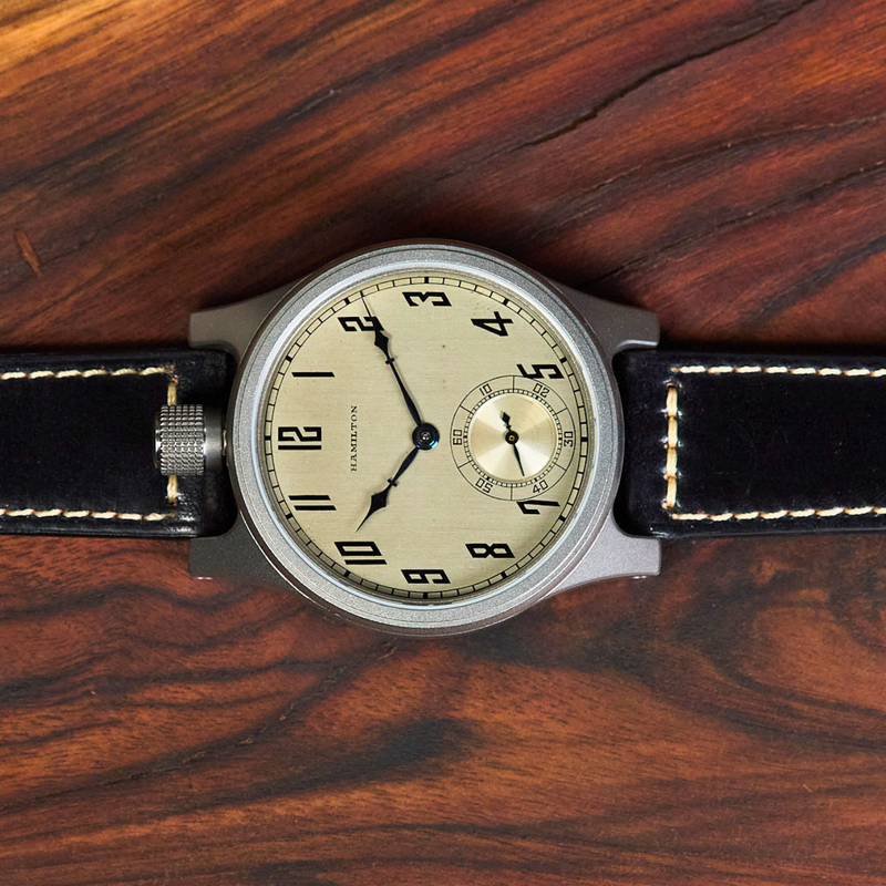 A wristwatch with a round silver-toned case, cream-colored dial, and black leather strap with white stitching, laid flat on a dark wood surface.