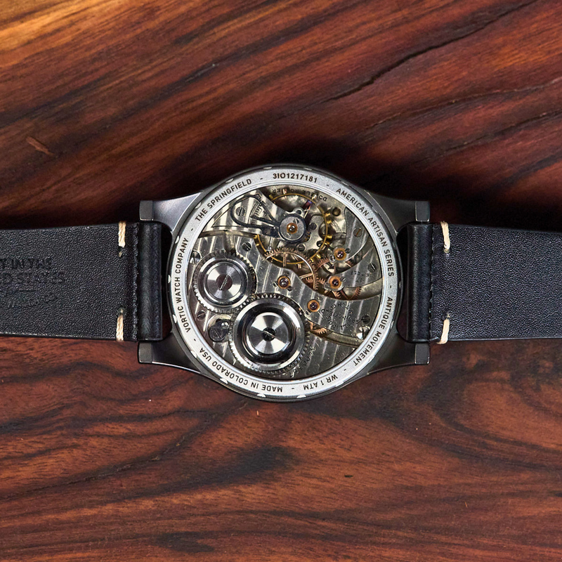 A close-up of a wristwatch laid flat on a dark wood surface, showing the open back case with visible mechanical gears and a black strap. The watch case appears dark gray to black, and the strap is black leather with small gold-toned hardware.