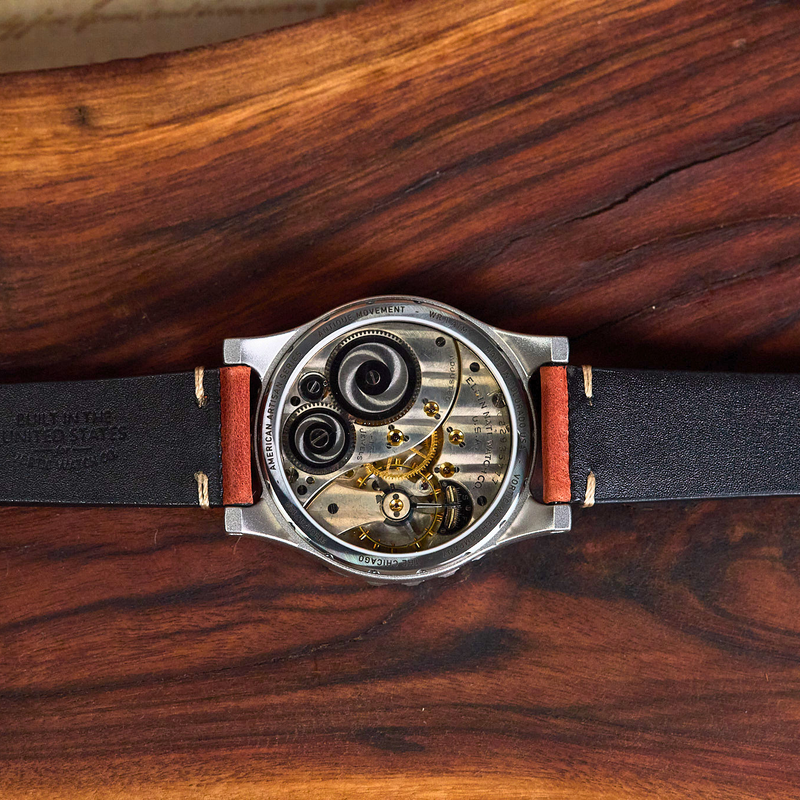 A close-up of a skeleton watch with a black leather strap and exposed mechanical movement, laid flat on a richly grained dark wood surface.