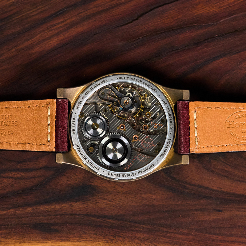 A close-up top-down view of a wristwatch with a transparent back showing the intricate mechanical movement, resting on a dark brown wooden surface. The watch has a tan leather strap with 'Built in the United States' and 'Horwatch' branding.