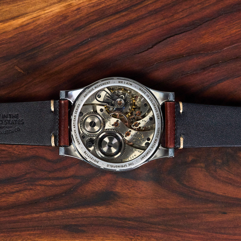 A close-up top-down view of a mechanical watch with a black leather strap and open back, showing the intricate steel and gold-toned movement, resting on a dark brown wood grain surface.