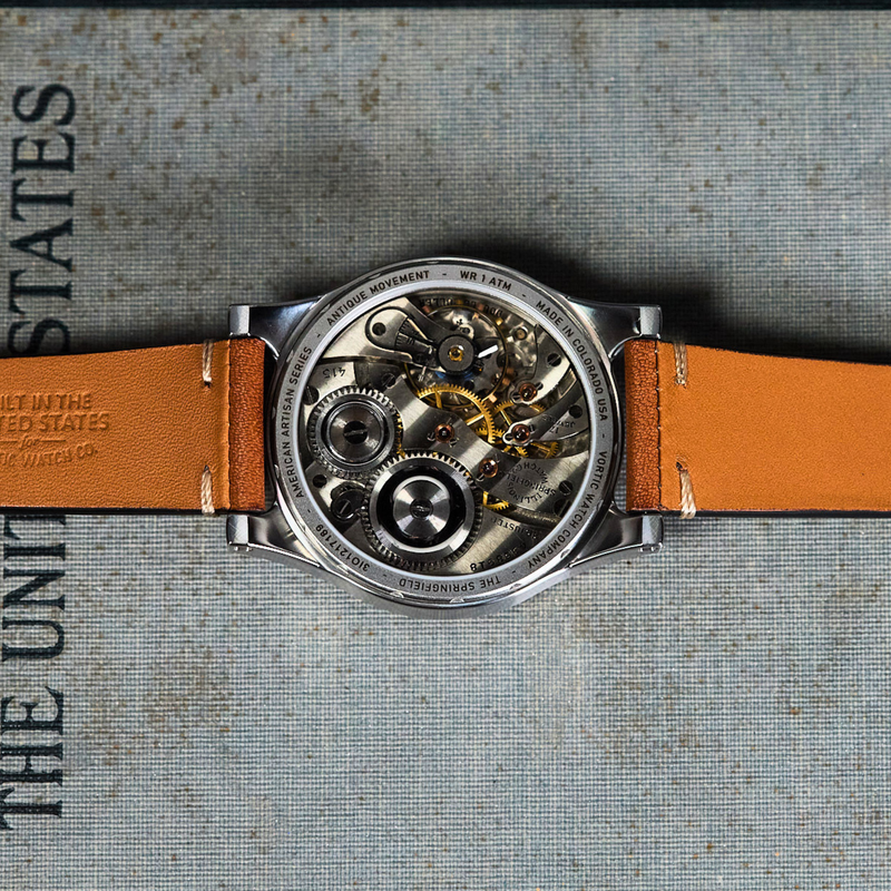 A close-up top-down view of a skeleton watch with a tan leather strap, showing the intricate mechanical movement through a clear case, laid flat on a textured gray surface with printed text.