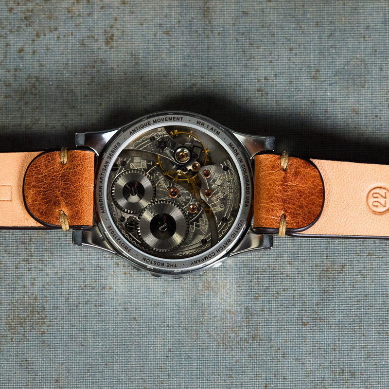A top-down view of a skeleton watch with a silver-toned case, showing the intricate mechanical movement through a clear dial, attached to a light tan leather strap with brown end caps, laid flat on a textured blue-gray background with printed text.