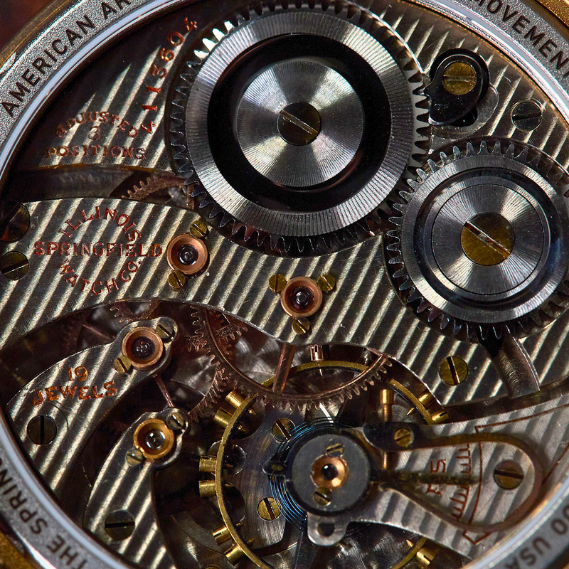 Close-up of a watch movement showing silver and gold-toned gears, screws, and plates, with engraved text around the edge and a reddish-brown leather strap visible at the sides.