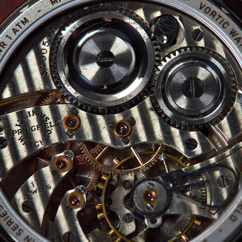 Close-up of a mechanical watch back showing intricate silver and gold-toned gears, screws, and plates, with engraved text including 'ANTIQUE MOVEMENT' and 'VORTEX WATCH COMPANY'.