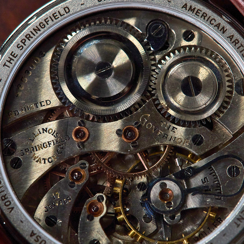 Close-up of a skeleton watch back showing the intricate mechanical movement, with engraved text including 'THE SPRINGFIELD' and 'ANTIQUE MOVEMENT', plus a brown leather strap visible at the edges.