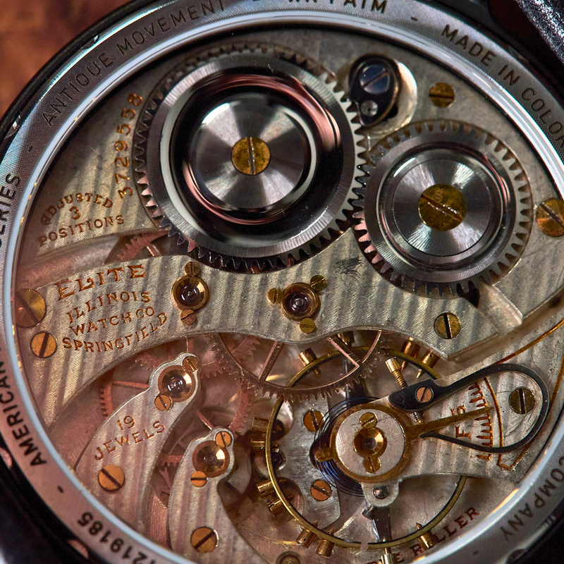 Close-up of a Vortic Watch Company Elite Automatic Movement watch showing the intricate mechanical movement with gold-toned gears, silver-toned components, and a black case on a leather strap.